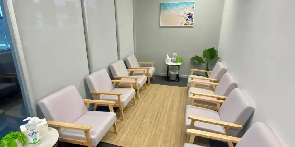 InspireHM— A small waiting room with six wooden chairs with gray cushions arranged along the walls. A light brown rug covers the floor. A round table with hand sanitizer and a plant is at the back, beneath a beach photo on a gray wall.