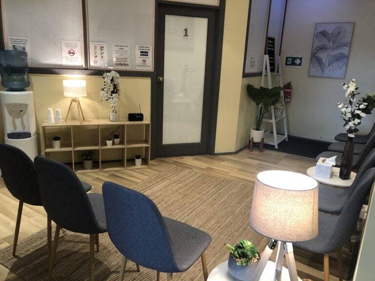 InspireHM— A waiting room with gray chairs arranged around a rug, a water cooler, decorative plants, and soft lighting from lamps. A door labeled "1" is in the background, and framed posters are on the walls.