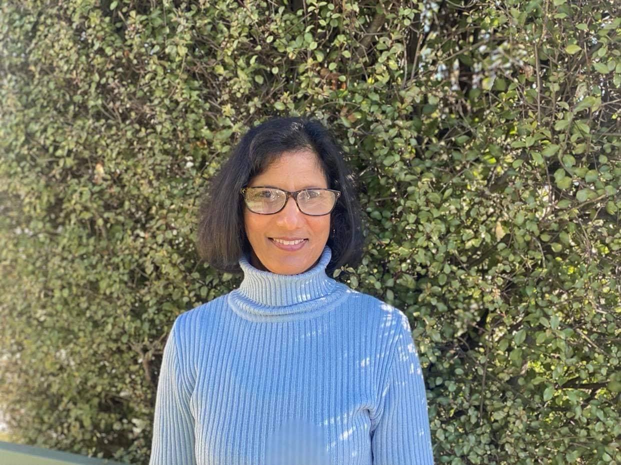 InspireHM— A person with glasses and a blue turtleneck sweater smiles in front of a lush green hedge on a sunny day.