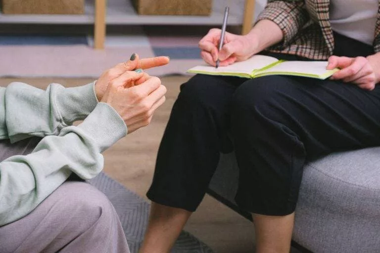 InspireHM— Two people are seated during a session. One person is taking notes in a notebook while holding a pen, wearing a plaid jacket. The other person is gesturing with hands, wearing a light green sweatshirt. Focus on hands and notebook.