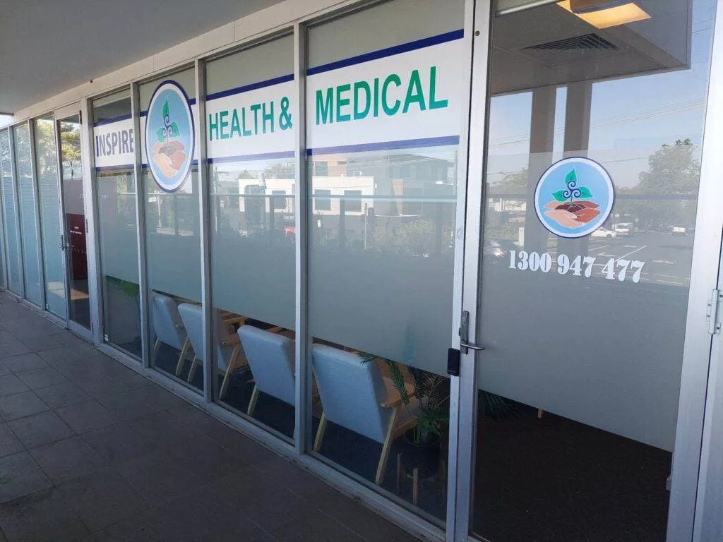 InspireHM— Exterior view of a health and medical clinic with frosted glass windows and a logo featuring a stethoscope and beach scene. The sign reads "Inspire Health & Medical" with a contact number. Several chairs are visible inside the entryway.
