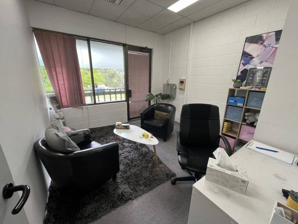 InspireHM— A small office with a window. Inside, there are two armchairs and a desk chair around a white coffee table on a gray rug. Shelves hold various items, and a painting hangs on the wall. The window has pink blinds partially opened.