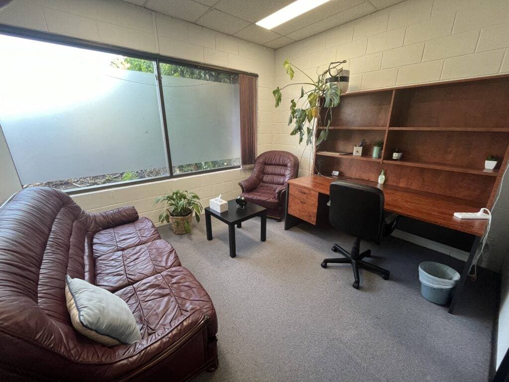 InspireHM— Office room with a brown leather couch and armchair, wooden desk with a black swivel chair, and a potted plant. The large window has frosted panels, and a shelf holds a few decorations. A tissue box and sanitizer sit on a small table.