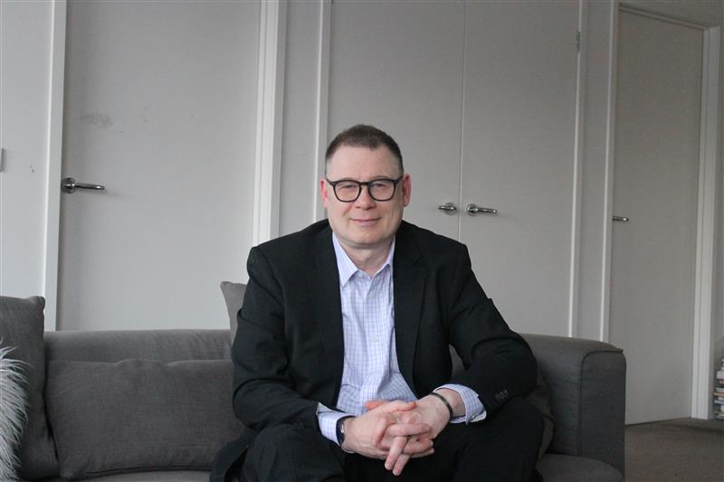 InspireHM— A man in a suit and glasses sits on a gray couch with his hands clasped, smiling slightly. The background shows a light-colored wall and closed doors. The setting appears to be an office or living space.