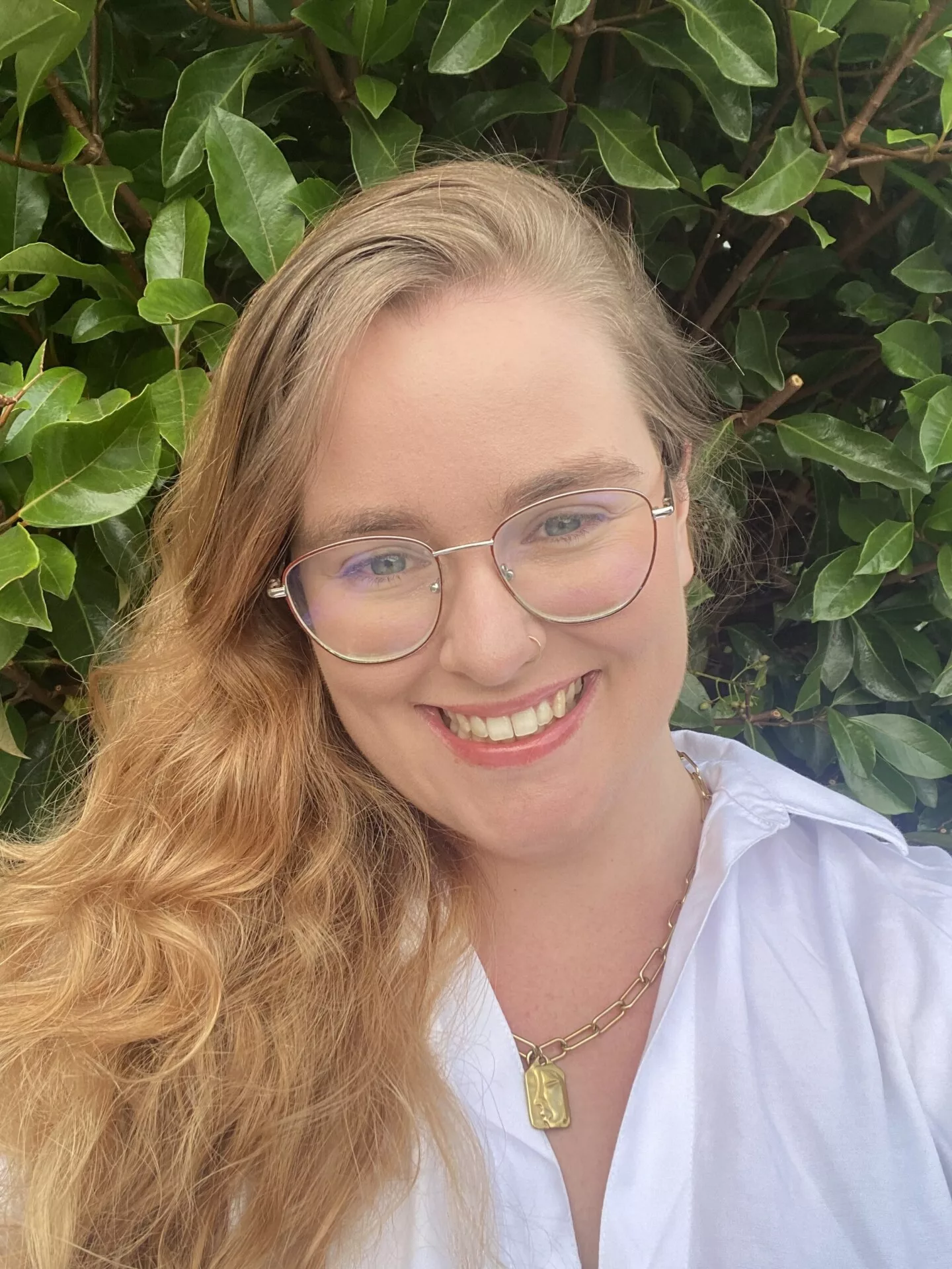 InspireHM— Person smiling with long, wavy blonde hair, wearing glasses, a white shirt, and a gold necklace. They are standing in front of lush green leaves.