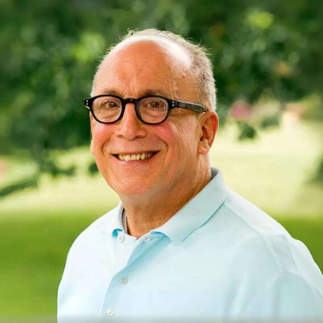 InspireHM— A smiling man wearing glasses and a light blue polo shirt is standing outdoors. The background is blurred with greenery and trees, suggesting a park or garden setting.