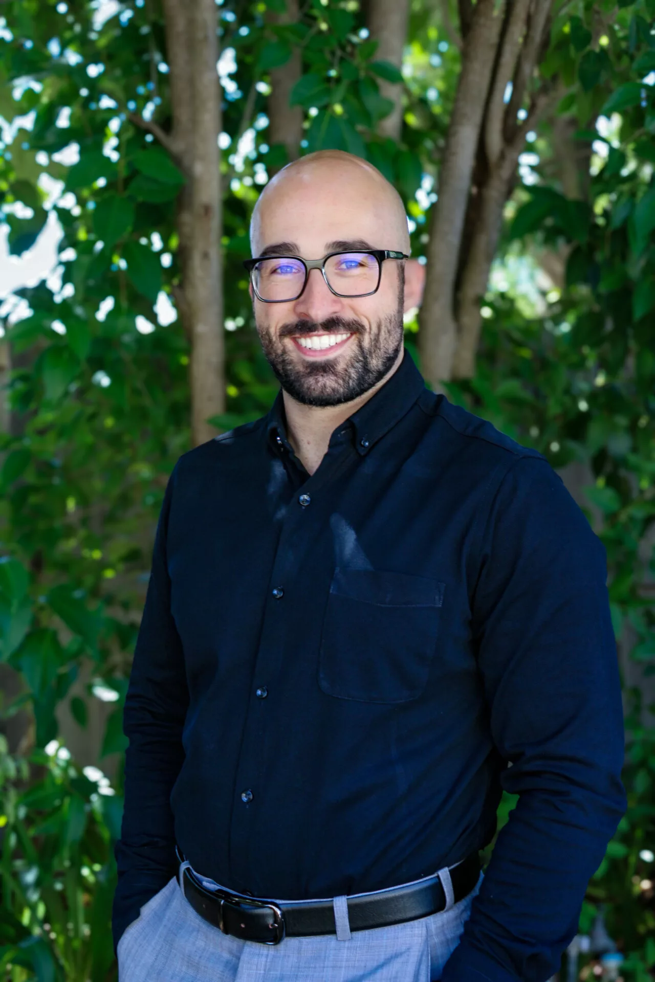 InspireHM— A person with a bald head, beard, and glasses is smiling while wearing a dark button-up shirt and light gray pants. They stand outdoors in front of lush green foliage.