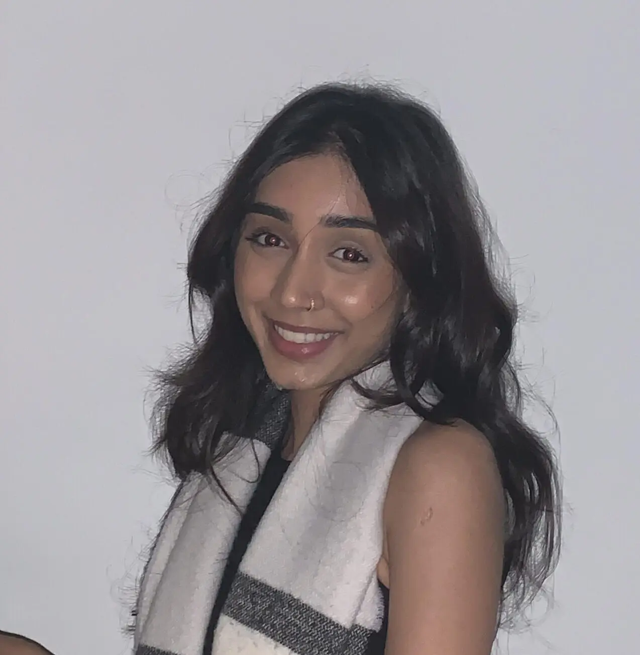 InspireHM— A young woman with long dark hair smiles at the camera. She is wearing a sleeveless top and a white scarf with gray stripes, standing in front of a plain light-colored wall.