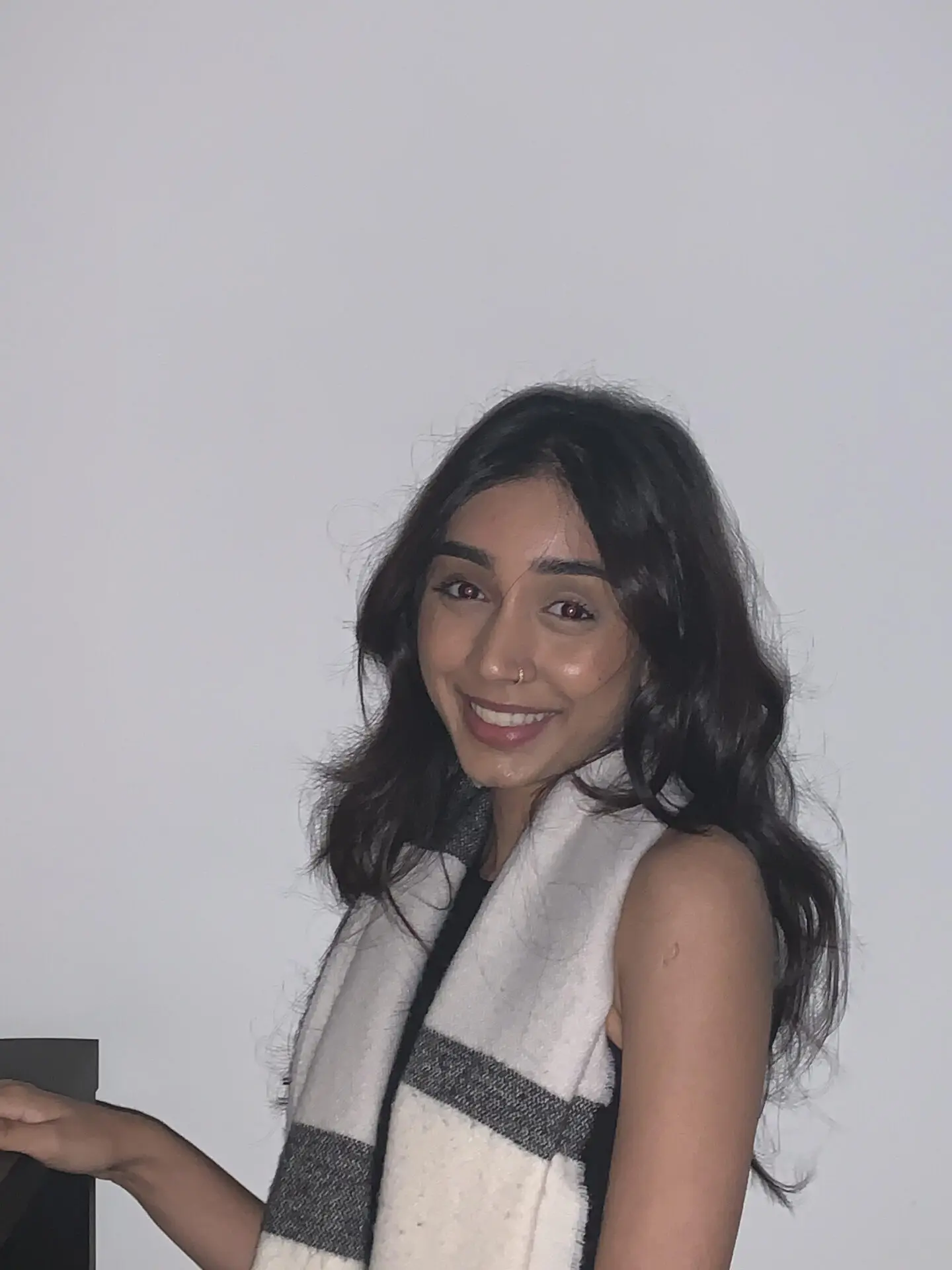 InspireHM— A young woman with long dark hair smiles at the camera. She is wearing a sleeveless top and a white scarf with gray stripes, standing in front of a plain light-colored wall.