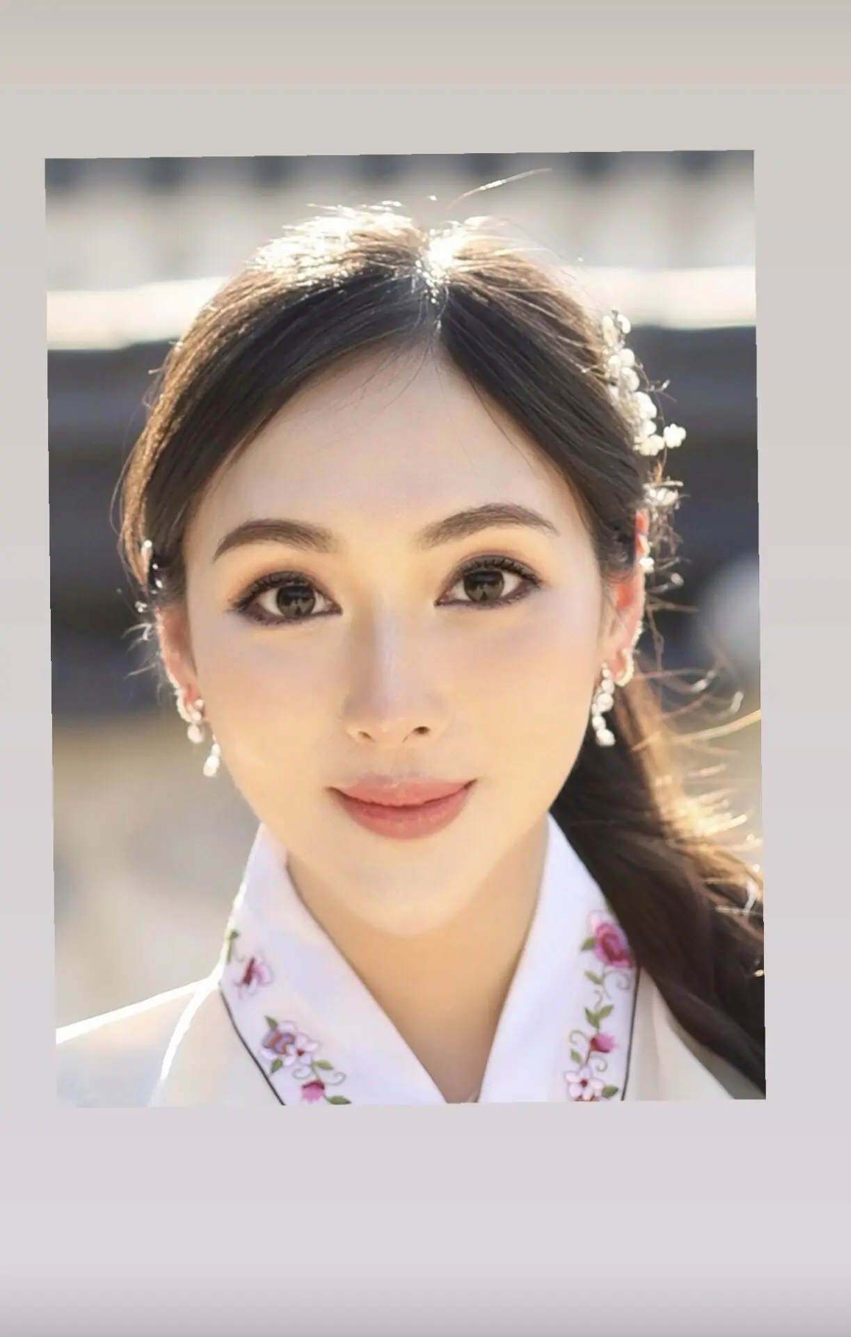 InspireHM— A young woman with long dark hair, wearing delicate earrings and a traditional white embroidered collar, smiles softly at the camera. She has natural makeup and floral hair accessories. The background is softly blurred.