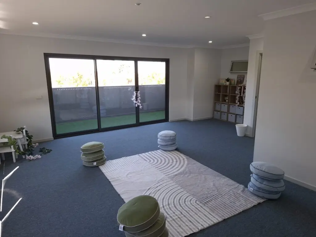 InspireHM— A bright, spacious room with blue carpet, a large rug, and round floor cushions arranged in a circle. There are sliding glass doors leading to a patio and cubby shelves with baskets against the wall. Plants and soft lighting create a calm atmosphere.