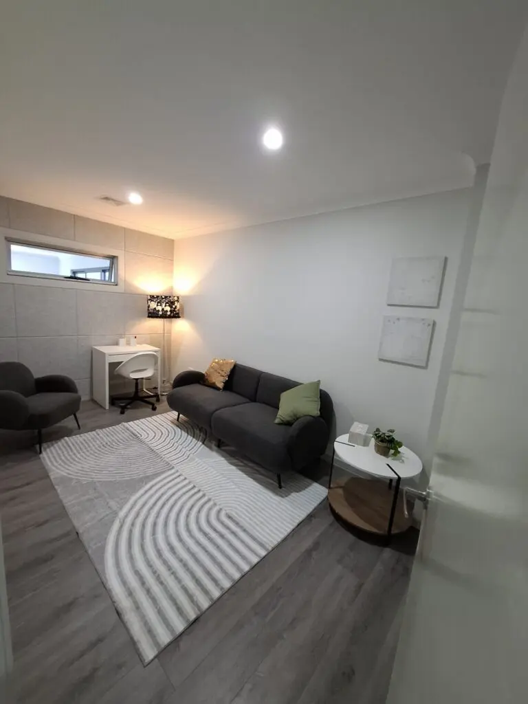 InspireHM— A modern, cozy office with a gray sofa, green and white cushions, a round side table, a patterned rug, a white desk with a chair, a black lamp, and two abstract wall art pieces on white walls.