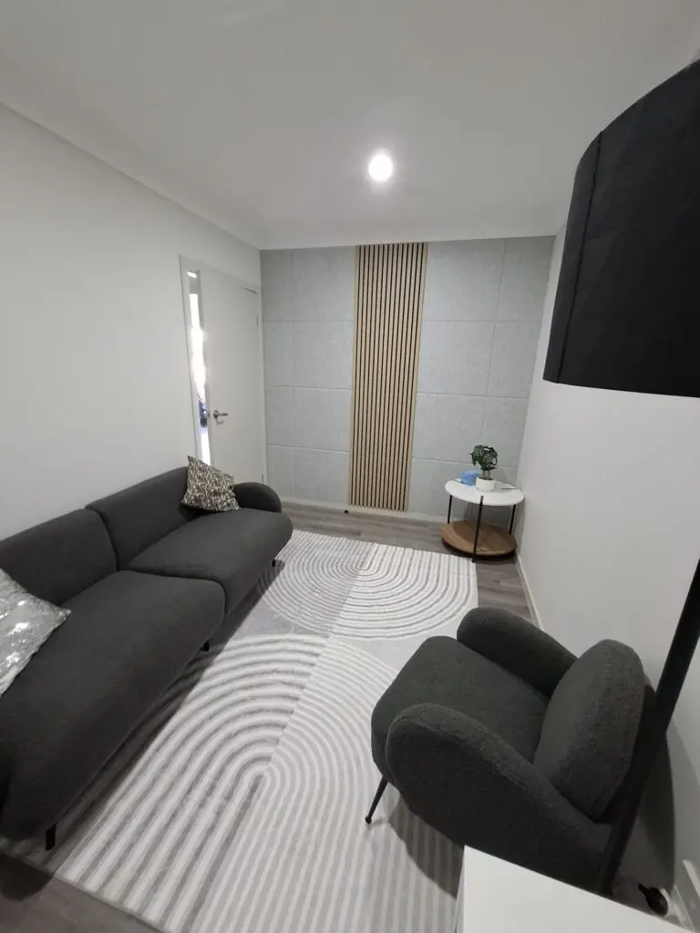 InspireHM— A modern, minimalist living room with a dark gray sofa and armchair, light patterned rug, small round side table with a plant, and a wooden slat feature on a gray accent wall.