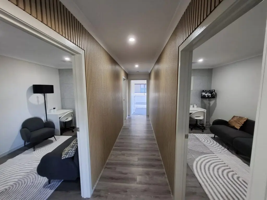 InspireHM— A modern hallway with light wood floors and vertical wood paneling, leading to a door. Two rooms on either side feature modern black sofas, white desks, and minimalistic decor.