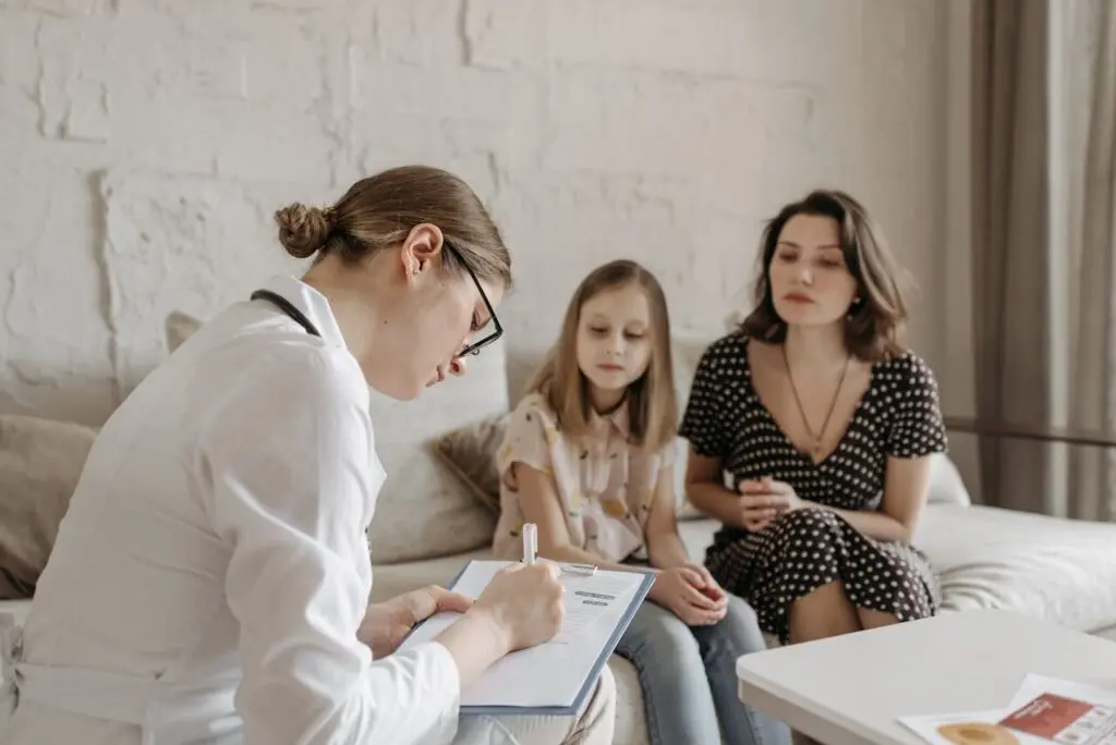 InspireHM— A doctor in a white coat writes on a clipboard while sitting across from a young girl and a woman, likely her mother, who are seated together on a couch in a bright room.