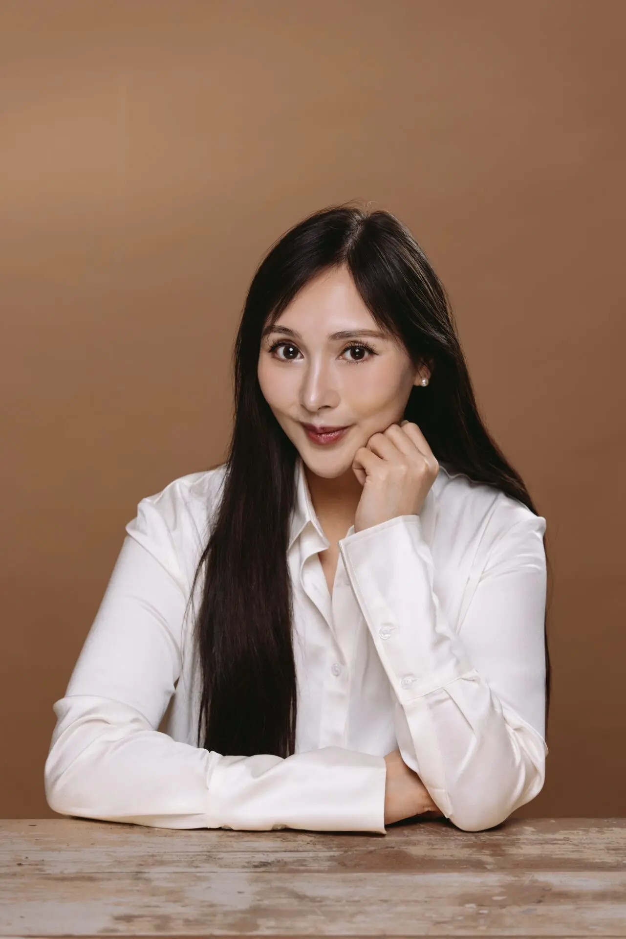 InspireHM— A woman with long dark hair, wearing a white button-up shirt, sits at a wooden table with her chin resting on her hand, smiling softly against a plain brown background.