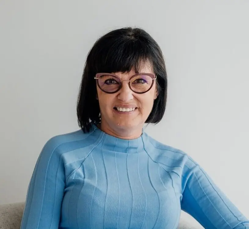 InspireHM— A woman with short dark hair and glasses, wearing a light blue ribbed sweater and light blue jeans, sits comfortably on a light-colored sofa against a plain white wall, smiling at the camera.