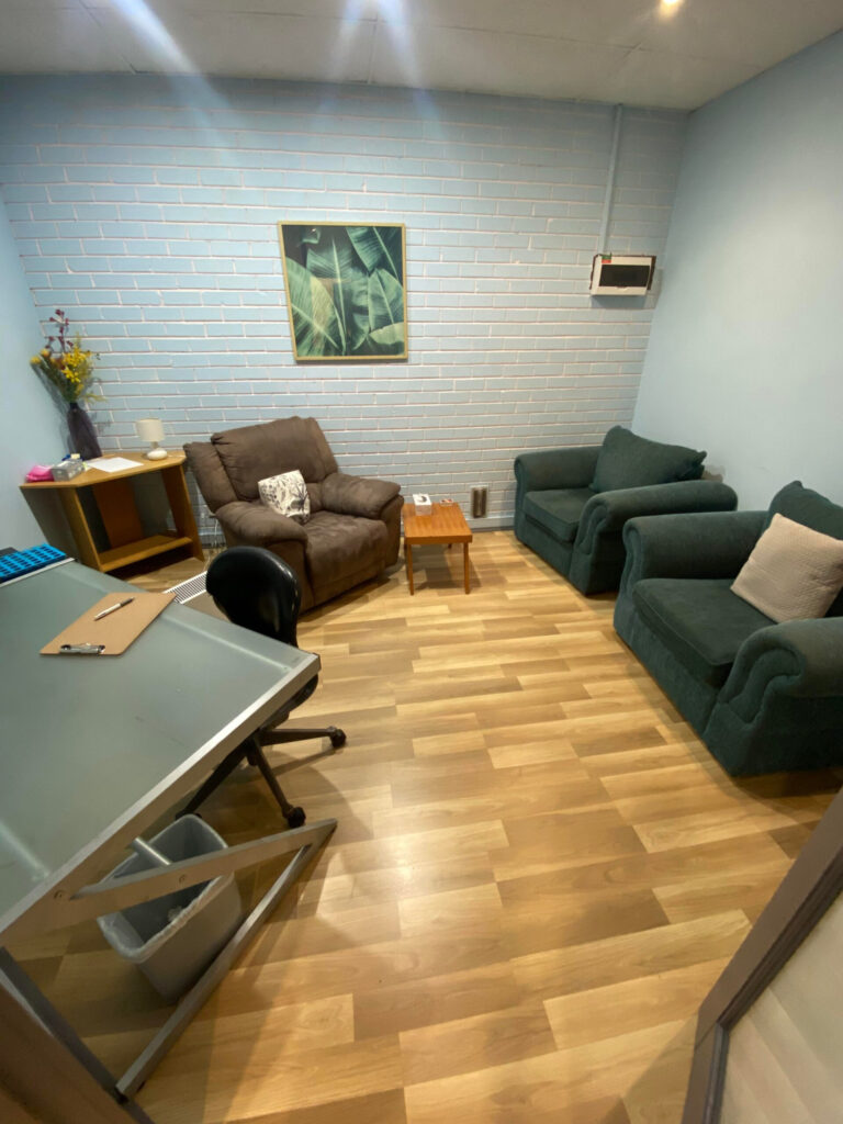 InspireHM— A small cozy office with light wood flooring, a glass desk with a black chair, a trash bin, two green armchairs, a brown recliner, a small wooden table, and a side table with a vase of flowers. A framed leafy print hangs on a blue brick wall.