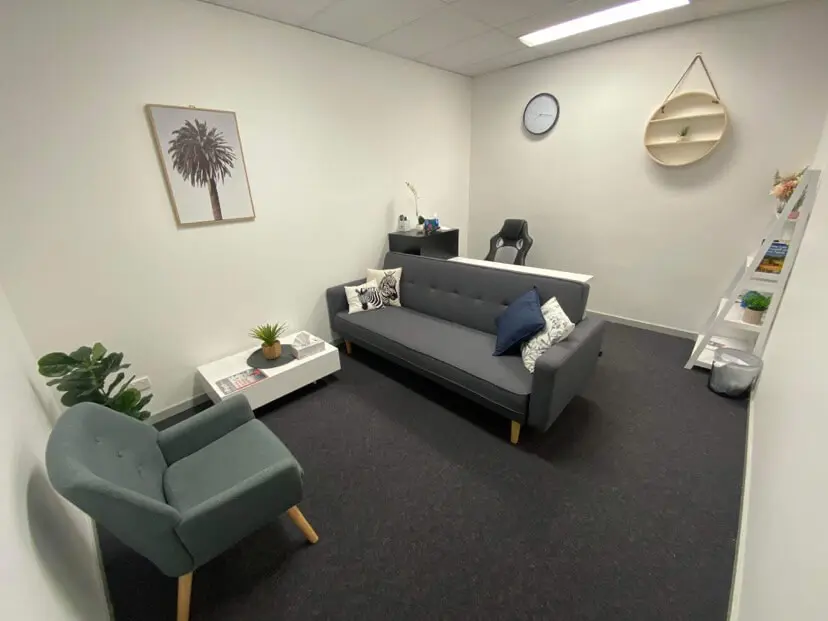 InspireHM— A modern office with white walls, a gray sofa with cushions, a gray armchair, a small white coffee table with a plant, a desk with a chair, a clock, a round shelf, a picture of a palm tree, and a ladder shelf with books and plants.