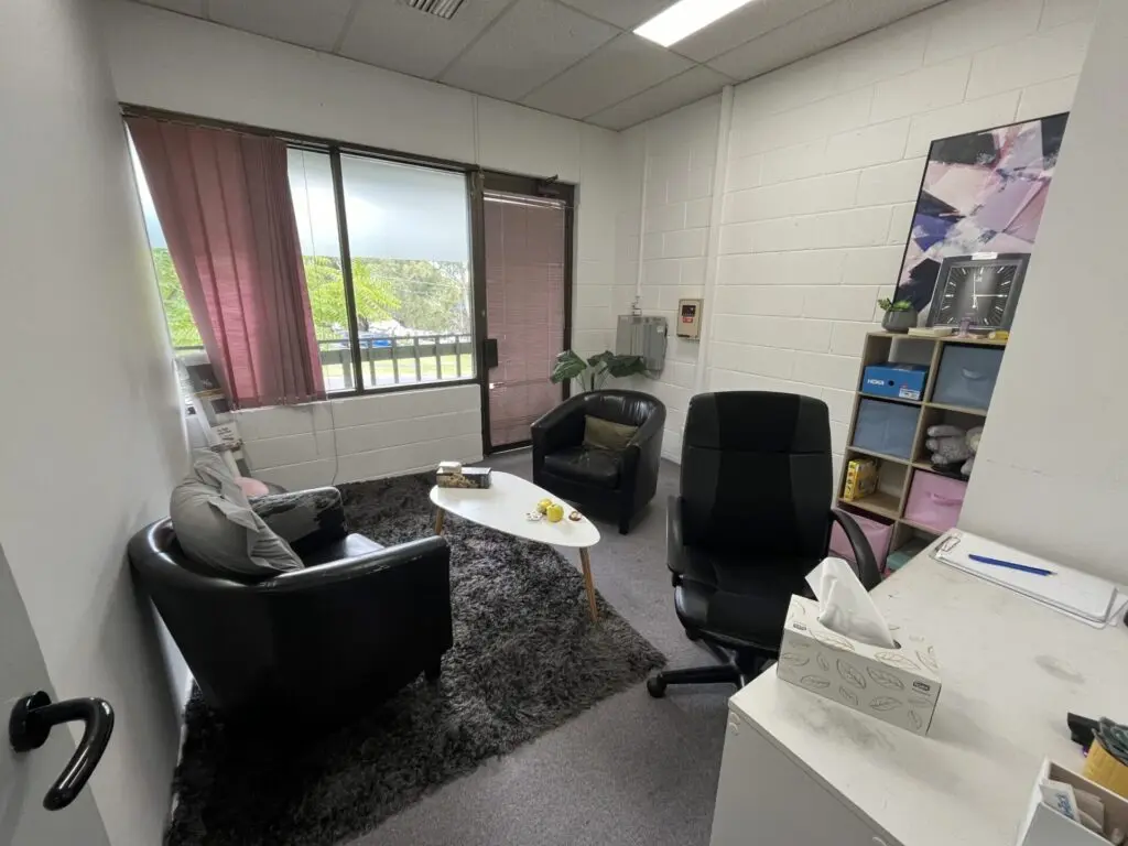InspireHM— A small office with two black chairs around a white coffee table on a shaggy rug, a desk with a black chair, tissue box, and some stationery, pink curtains partially cover a window, and shelves with books and decorations are in the corner.